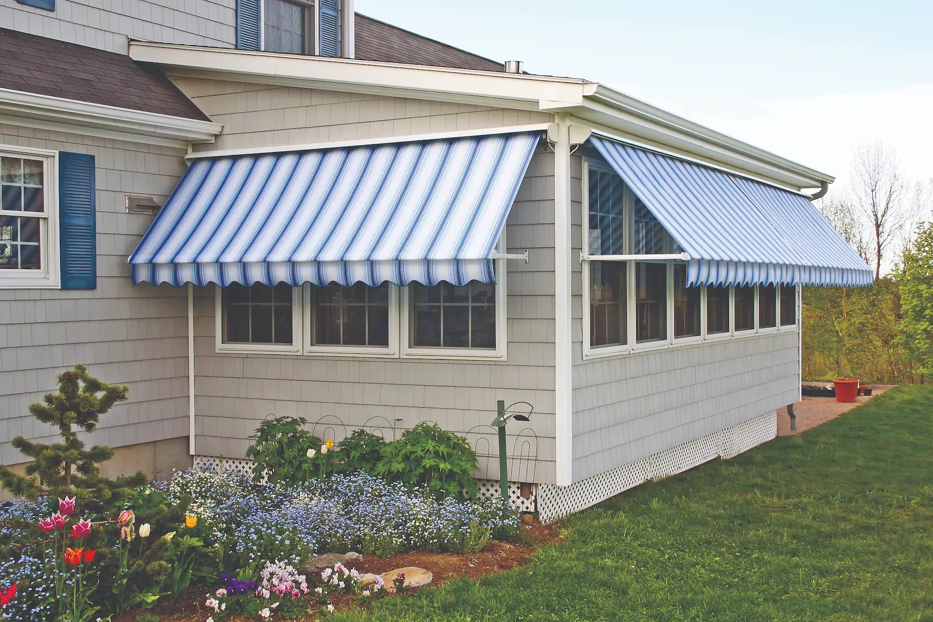 Exterior shot of a sunroom with windows shaded by a blue-and-white-striped retractable awning. Color and pattern choice come into play when investigating Fixed vs. Retractable Window Awnings.