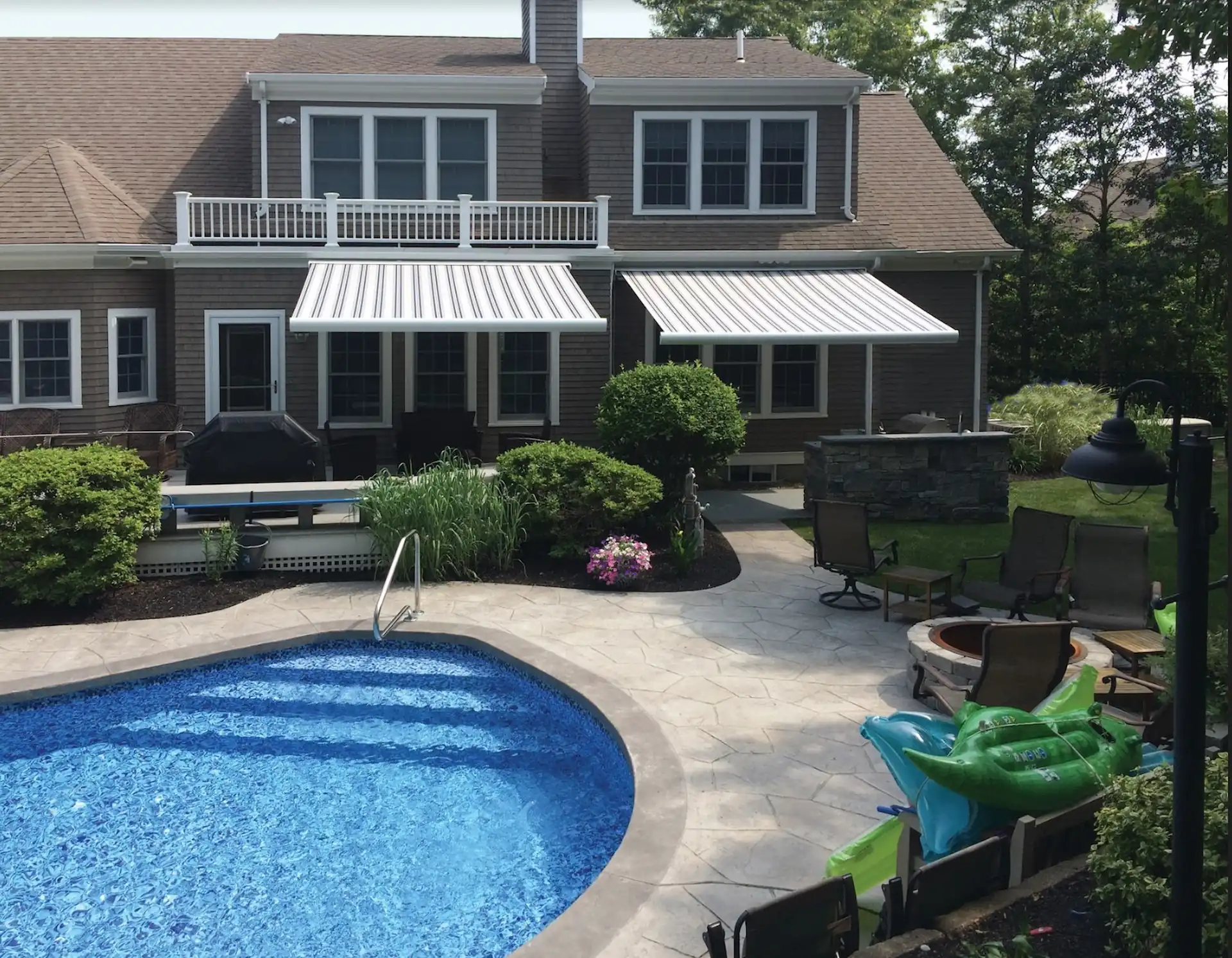 Exterior shot of a pool deck situated next to a stylish, brown house with 2 striped roll up awnings extended over the patio.